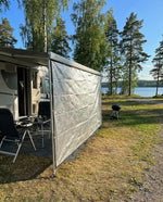 Ljust sol- och insynsskydd uppspänt längs husvagnens sida vid en sjö med björkar – skyddar mot sol och insyn