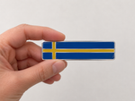 Hand som håller en avlång självhäftande svensk flagga i emalj – dekal för husbil och husvagn